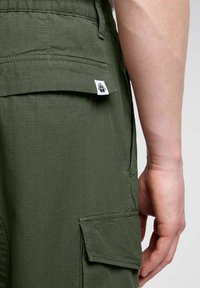 Pantaloni cargo verdi realizzati in tessuto leggero, dotati di una tasca posteriore e di un'etichetta con un logo grafico sulla vita.
