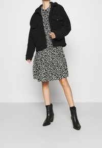 Giacca nera e morbida con due tasche frontali sopra un vestito floreale nero. Il vestito presenta piccoli fiori bianchi e una gonna a balze. Stivaletti neri lucidi.