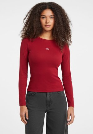 Maglietta rossa a maniche lunghe realizzata in tessuto a costine, caratterizzata da una vestibilità slim e un piccolo logo bianco "Guess Jeans" sul petto. Abbinata a pantaloni grigio scuro.