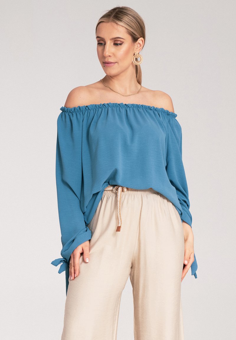 Bluzka z odkrytymi ramionami w turkusowym kolorze z marszczonym dekoltem, długimi rękawami z wiązaniami, w zestawie z beżowymi spodniami szerokimi w nogawkach.