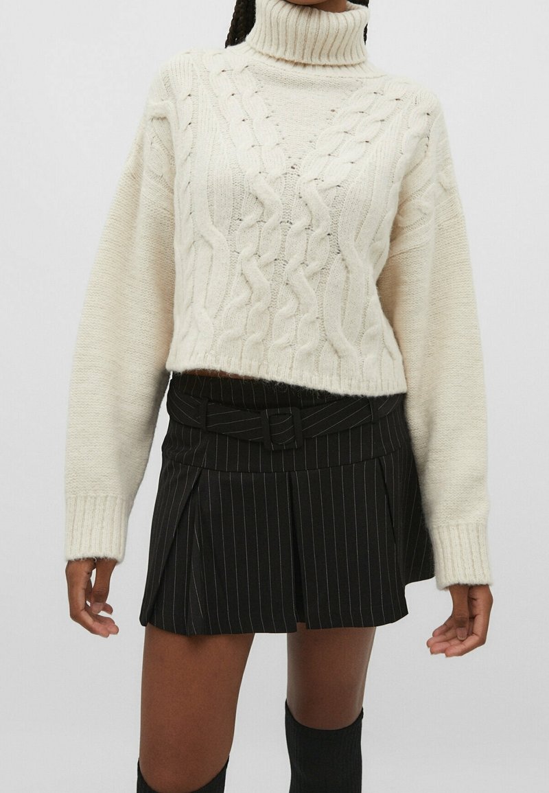 Femme portant un pull à col roulé tricoté torsadé couleur crème et une mini-jupe plissée à fines rayures noires avec une ceinture assortie, visible des épaules aux cuisses.