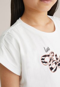 Balta medvilninė marškinė su trumpomis pūstomis rankovėmis, puošta juodais ir oranžiniais siuvinėtais užrašais bei dekoratyviniu piešiniu. Minkšta tekstūra.