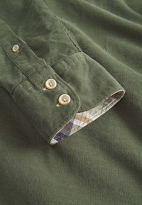 Cuff di camicia in corduroy verde con due bottoni chiari e un motivo a quadretti a contrasto all'interno del polsino.