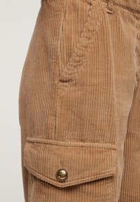 Bruine ribfluwelen cargobroek heeft verticale ribbels, zijzakken en een knoopsluiting op de cargozak. De gestructureerde stof biedt een zachte afwerking.