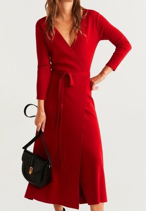 Femme portant une robe portefeuille rouge à manches longues avec une ceinture à la taille, tenant un sac à main noir par sa lanière dans sa main gauche.