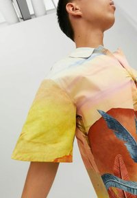 Camicia a maniche corte in tessuto leggero. Presenta una vivace sfumatura di giallo e rosa con motivi di foglie tropicali in blu e rosso.