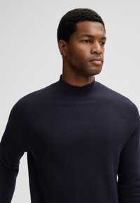 Junger Mann mit kurzen lockigen Haaren, der einen navyblauen, gerippten Pullover mit Stehkragen und einem kleinen Ohrstecker trägt und zur Seite schaut.