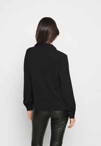 Camicetta nera a maniche lunghe in tessuto morbido, con colletto e polsini abbottonati, indossata con pantaloni in pelle nera aderenti.