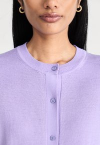 Lavendelstickad kofta med rund krage och fyra matchande knappar mitt fram. Slät textur och figurnära design.