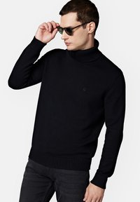 Czarny sweter z golfem, wykonany z dzianiny, z ryflowanymi mankietami i dołem. Model, nosząc okulary przeciwsłoneczne, pozuje z ręką na okularach.