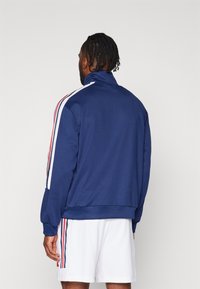 Giacca a zip blu navy con colletto alto, dotata di strisce laterali bianche e rosse, realizzata in tessuto liscio, indossata sopra pantaloni corti bianchi.