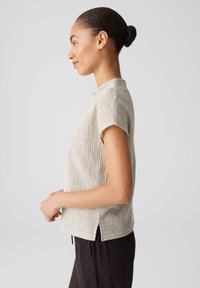 Short-sleeve, textured light beige top with a high neck and side slits, paired with dark pleated trousers.