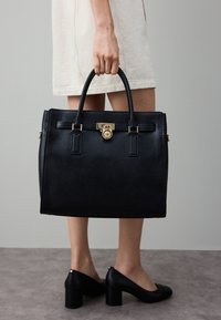 Zwarte leren handtas met gestructureerd ontwerp, gouden beslag, bovenhandvatten en een slotdetail. Gestructureerd oppervlak en rechthoekige vorm.