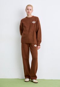 Brauner Strickpullover und passende Hose mit weißen Adidas-Logoakzenten. Weicher Stoff, lockere Passform und gerippte Bündchen an Ärmeln und Saum.