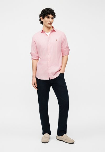 Άνδρας στέκεται με το αριστερό χέρι στην τσέπη, φορώντας ροζ πουκάμισο με κουμπιά, σκούρο παντελόνι και μπεζ μοκασίνια, πάνω σε απλό λευκό φόντο.