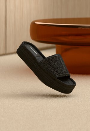 Sandale à plateforme noire tissée avec marquage "Calvin Klein" sur la semelle intérieure et la tige, présentée en suspension sur un fond neutre et une table marron.