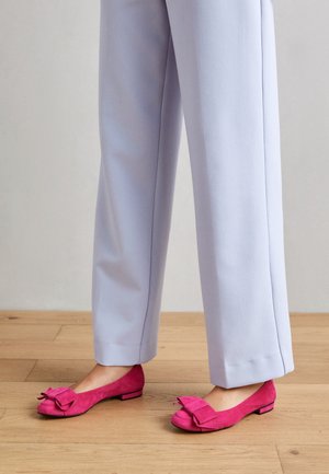 Ballerines en daim rose avec un nœud décoratif, semelle plate et bout arrondi, associées à un pantalon gris clair à jambes larges sur un sol en bois.