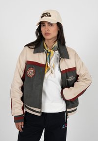 Femme portant une veste color block beige, grise et rouge, une écharpe à motif, une casquette beige avec texte, une chemise blanche et un pantalon noir avec logo.