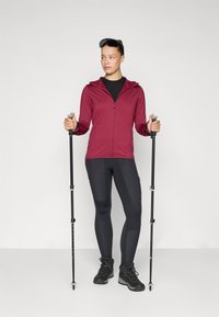 Bordeauxfarbener Reißverschluss-Hoodie, schwarzes enges Oberteil, schwarze Leggings mit Kniefelgen, schwarze Wanderschuhe und Trekkingstöcke. Einfarbiger Hintergrund.