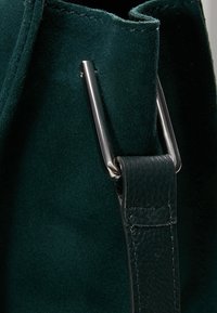 Vihreä samettinen käsilaukku, jossa on sileä nahkahihna ja hopea tarvikkeet. Yksityiskohta näyttää laukun reunan ja hihnan kiinnityksen.