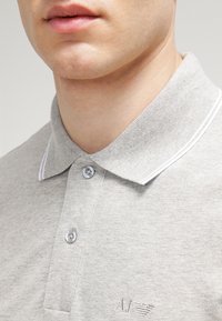 Šedé polo tričko s texturovanou tkaninou, dvěma průhlednými knoflíky a bílým límcem s lemováním. Na levé straně má malé logo.