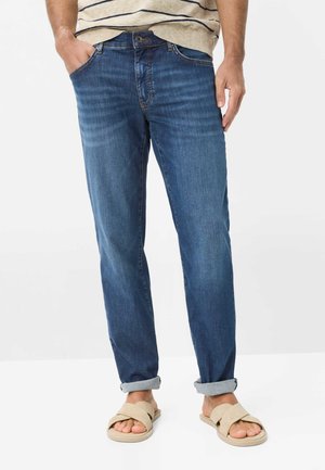 Mand iført opklebede blå jeans, beige stribet sweater og beige slip-on sandaler står med hænderne i lommerne.