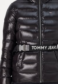 Svart pufferjakke med blank overflate, horisontal søm, glidelås foran og belte med merkevaren "TOMMY JEANS" i hvit skrift.