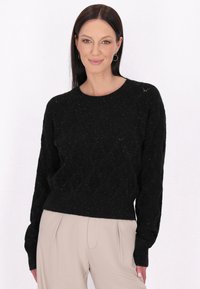 Pulover negru texturat cu un model în diamant, decolteu rotund și manșete cu margine ondulată. Asortat cu pantaloni bej deschis.