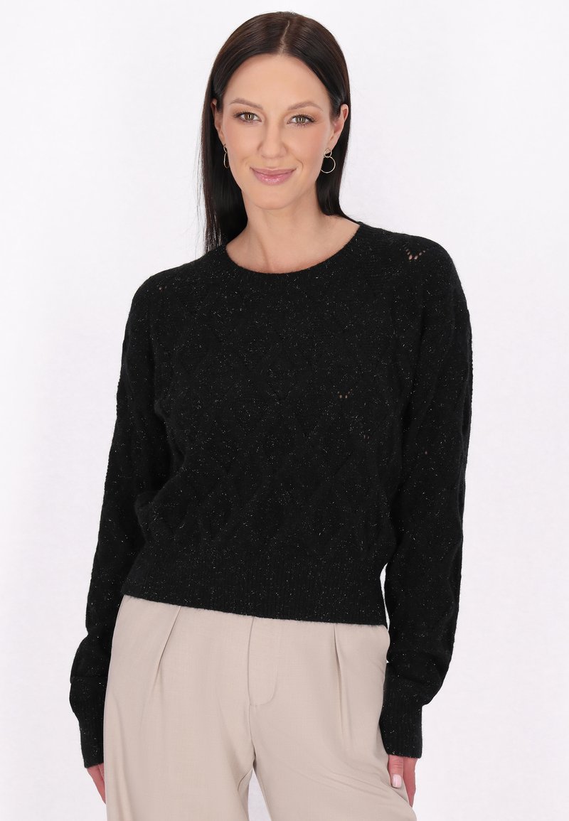 Pulover negru texturat cu un model în diamant, decolteu rotund și manșete cu margine ondulată. Asortat cu pantaloni bej deschis.