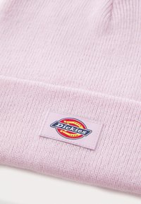Lyserød strikket hue lavet af blødt stof, med et mærke med "Dickies"-logoet i blå og rød, syet på en matchende lap.