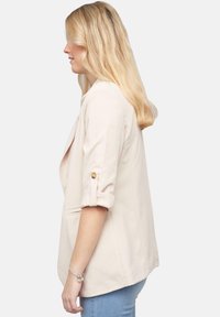 Lichtbeige blazer met opgerolde mouwen en gouden knoopaccenten. Heeft een losse pasvorm en twee zijzakken. Zachte textuur, minimalistisch ontwerp.