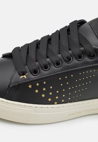 Černé kožené tenisky s černými tkaničkami, zlatými hvězdnými akcenty a texturovanou bílou podrážkou s brandingem. Hvězdy se liší velikostí a umístěním.