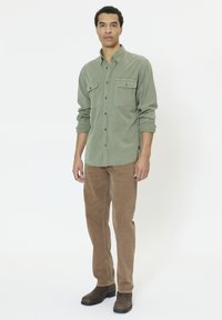 Mees seisab valge tausta ees, kandes rohelist nööbitavat särki, pruune pükse ja tumedat pruuni kingi, käed lõdvestunult külgedel.