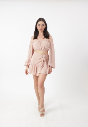 Off-shoulder, lichtroze croptop met lange mouwen; bijpassende wikkelrok met ruches aan de zoom. Gedragen met sandalen met bandjes.