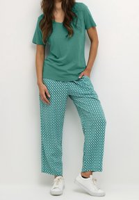 Tyrkysové tričko s véčkovým výstřihem spárované s širokými kalhotami s potiskem v tyrkysové a bílé barvě. Model má na sobě bílé tenisky, ruce v kapsách.