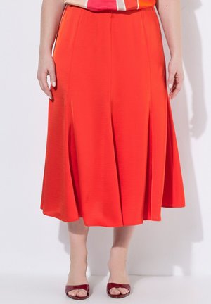 Falda de longitud media de color rojo brillante con pliegues suaves, combinada con tacones burdeos de punta abierta sobre un fondo blanco liso.
