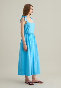 Lichtblauwe katoenen jurk met vierkante halslijn, verstelbare strikbandjes, elastische taille en een volle rok. Gecombineerd met meerkleurige platte sandalen.