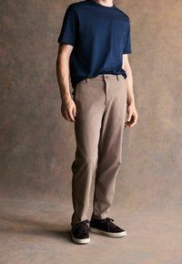 Jūras spēku apaļā kakla t-kreklu, bēša taisna griezuma bikses un tumši sporta apavi, stāvot pretī neitrālam teksturētam fona.