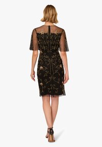 Žena v černých šatech ke kolenům s zlatou květinovou výšivkou a průhlednými krátkými rukávy, viděná z pohledu zezadu, v černých vysokých podpatcích.