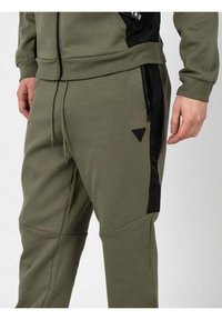 Pantalones de chándal verde oliva fabricados en un tejido suave, con un panel lateral negro, bolsillos con cremallera y un logotipo triangular en la pierna izquierda.