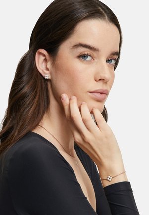 Die Ohrringe bestehen aus gruppierten weißen Perlen auf einer silbernen Basis. Das Model trägt ein schwarzes Oberteil und ein zartes Armband mit ähnlichem Perlenmuster.