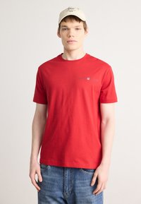 T-shirt en coton rouge à manches courtes et col rond ; arbore un petit logo gris sur la poitrine. Porté avec un jean en denim bleu.