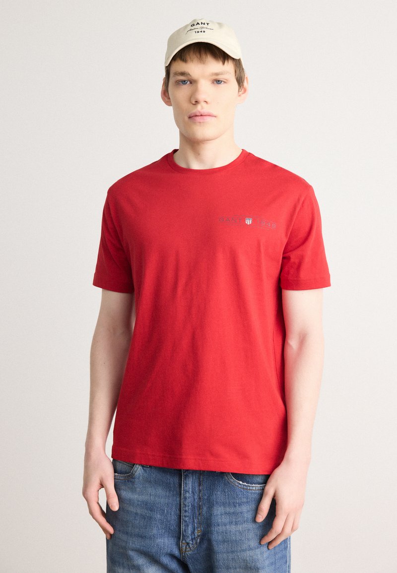 T-shirt en coton rouge à manches courtes et col rond ; arbore un petit logo gris sur la poitrine. Porté avec un jean en denim bleu.