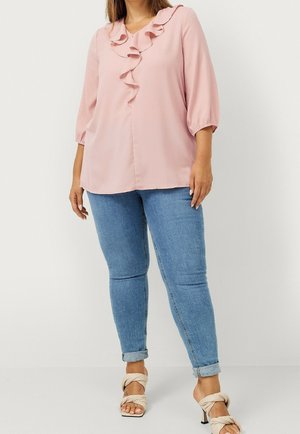 Femme portant un chemisier rose à volants avec des manches trois-quarts, un jean skinny bleu clair retroussé et des sandales beiges à talons.