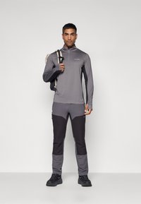 Maglia grigia a maniche lunghe con zip a 1/2 e logo, abbinata a pantaloni neri e grigi con tasche zippate e design a blocchi di colore.