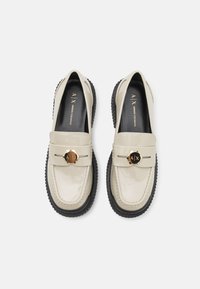 Beige lakleren loafers met zwarte rubberen zool, voorzien van een gouden logo accent en verfijnde stiksels rond de randen.