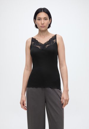 Žena s krátkymi tmavými vlasmi, oblečená v čiernej tielkovej blúzke s čipkovaným lemom a voľných sivých nohaviciach, stojaca pred jednoduchým pozadím.