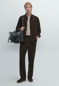 Dunkelbraune Knopfleiste über einem beigen Oberteil, braune Hose, schwarze Tasche und dunkle Schuhe. Glattes Material, lässige Passform und minimalistisches Design.