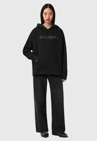 Fekete kapucnis pulóver "ALLSAINTS" logóval, elülső zsebével és hosszú ujjú kialakítással, széles szárú fekete nadrággal és lapos cipővel párosítva.