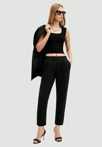 Top cropped preto combinado com calças pretas de cintura alta. A modelo segura um blazer preto, usando óculos de sol escuros e sandálias de salto pretas.